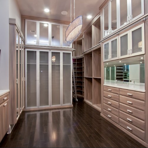 Elegant Luxury Walk-In Closet With Glass-Front Cabinets, Dark Hardwood Flooring, And Modern Chandelier Lighting.